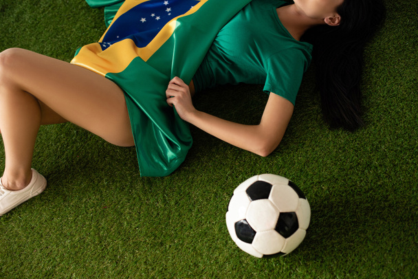 καλλιεργημένη άποψη της σέξι γυναίκα ξαπλωμένη με μπάλα ποδοσφαίρου και βραζιλιάνικη σημαία στο πράσινο γρασίδι - Φωτογραφία, εικόνα