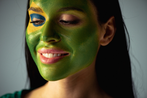 smiling female football fan with painted brazilian flag on face isolated on grey - Photo, Image