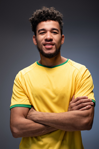 szczęśliwy amerykański fan piłki nożnej w żółtej koszulce stojącej z skrzyżowanymi ramionami na szarości - Zdjęcie, obraz