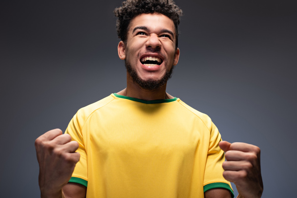 vzrušený africký americký fotbalový fanoušek ve žlutém tričku křičící a gestikulující na šedi - Fotografie, Obrázek
