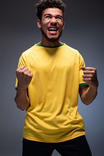 emocionální africký americký fotbalový fanoušek ve žlutém tričku křičící a gestikulující na šedi - Fotografie, Obrázek