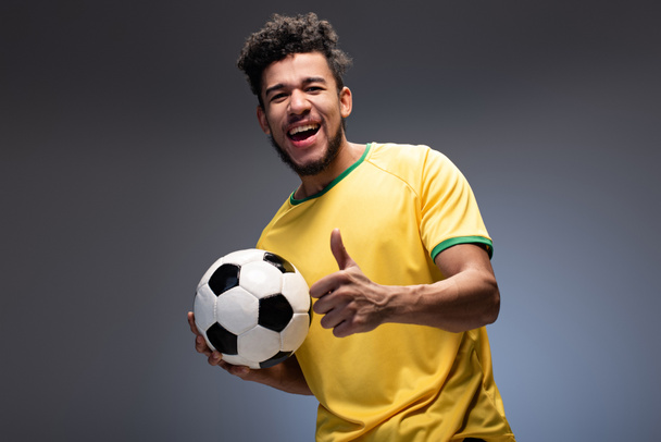happy african american football fan in yellow t-shirt holding ball and showing thumb up on grey - Фото, изображение