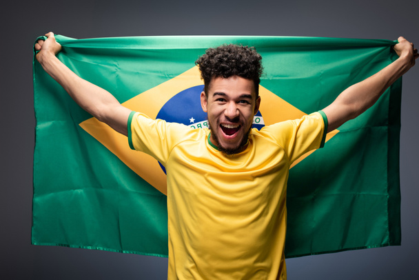 excited african american football fan screaming and holding brazilian flag on grey - 写真・画像