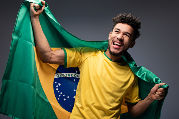 szczęśliwy african amerykański kibic piłki nożnej gospodarstwa brazylijska flaga na szary - Zdjęcie, obraz