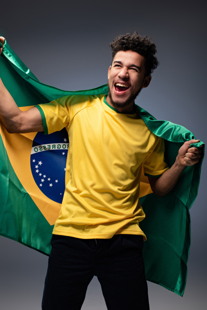 excited african american football fan holding brazilian flag on grey - Foto, afbeelding