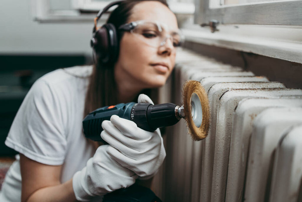 Beautiful and diligent middle age handy woman renovating her old home or apartment while listening to music with headphones. She is holding professional grinder and brushing radiator for later painting. Do it yourself housework concept. - Foto, Imagen