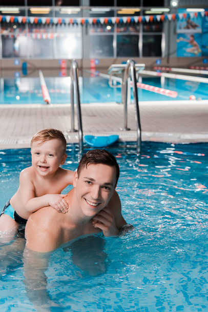 χαρούμενο παιδάκι και προπονητής κολύμβησης κοιτάζοντας την κάμερα στην πισίνα  - Φωτογραφία, εικόνα