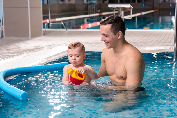 水泳プールで遊んでいる幼児の男の子を見ている間にハンサムな水泳トレーナーの笑顔  - 写真・画像