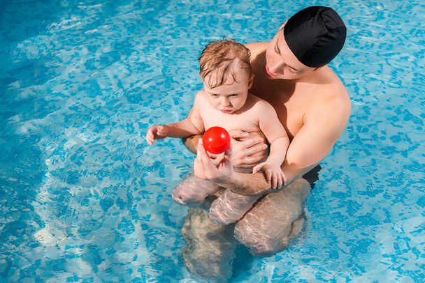 κολύμπι προπονητής στην πισίνα καπέλο κρατώντας μπάλα κοντά χαριτωμένο μικρό παιδί στην πισίνα  - Φωτογραφία, εικόνα