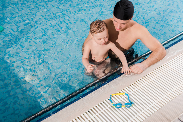 vista aérea del entrenador de natación en gorra de natación y niño pequeño mirando gafas
  - Foto, Imagen