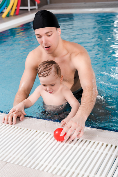 handsome swim coach in swimming cap touching ball near cute toddler boy in swimming pool  - Photo, Image
