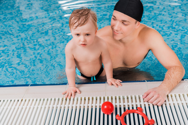 apuesto entrenador de natación en gorra de natación mirando niño en la piscina
  - Foto, Imagen
