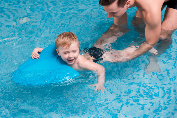 興奮した幼児は水泳コーチの近くでフラッターボードで泳ぐ  - 写真・画像