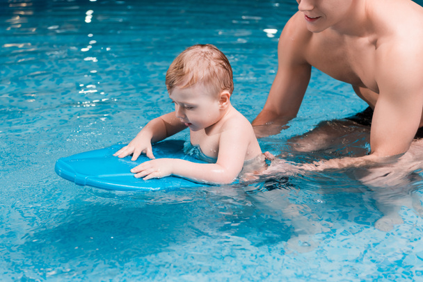 csecsemő fiú úszás lengődeszkával az izmos úszóedző közelében  - Fotó, kép