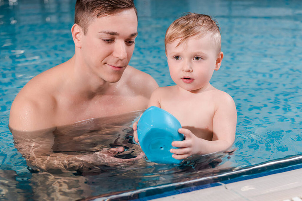 swim coach looking at toy ship in hands of toddler boy in swimming pool  - Photo, Image