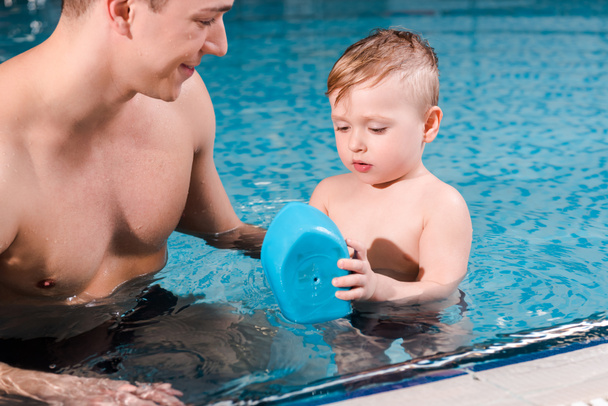 щасливий тренер з плавання, дивлячись на хлопчика, який грає з іграшковим кораблем у басейні
  - Фото, зображення
