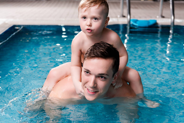 Yakışıklı yüzme koçu yüzme havuzunda kucağında küçük tatlı bir çocuk tutuyor.  - Fotoğraf, Görsel