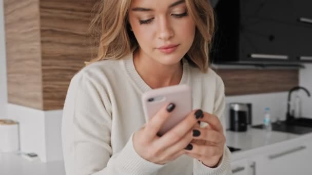 A pleased young blonde woman is using her phone in the kitchen in the morning - Filmmaterial, Video