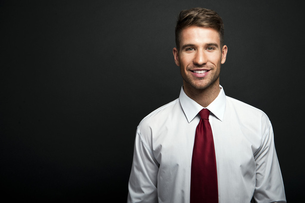 Retrato de perfil de un joven empresario sonriente y confiado, vestido con traje y corbata, frente a una pared oscura con espacio para copias
 - Foto, Imagen