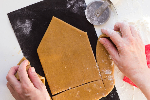 Woman hands removing excess raw gingerbread dough after cutting out wall of gingerbread house on black teflon baking sheet on white background - Foto, Bild