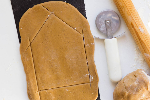 Cutting out wall of gingerbread house with knife for pizza on rolled raw gingerbread dough teflon baking sheet on the white background - Foto, Imagem