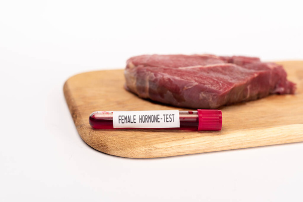 Foco seletivo do tubo de teste com teste de hormônio feminino em tábua de corte com carne crua isolada em branco
 - Foto, Imagem