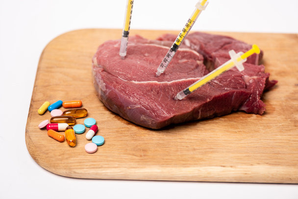 Strzykawki w surowym mięsie i tabletki hormonalne na desce do krojenia izolowane na białym - Zdjęcie, obraz