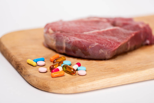 Selective focus of hormonal pills and raw meat on wooden cutting board isolated on white - Photo, Image
