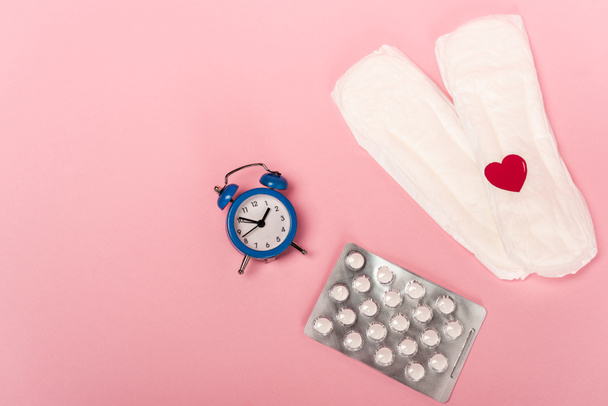 Bovenaanzicht van vrouwelijke pads met papieren hart, anticonceptiepillen en wekker op roze achtergrond - Foto, afbeelding