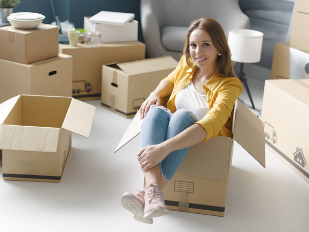 Smiling young woman moving in her new home, she is sitting in a cardboard box and smiling - Foto, imagen