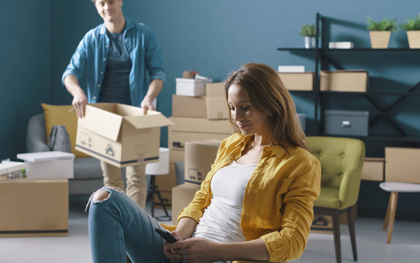 Woman sitting on a chair chatting with her phone during home relocation, her boyfriend is moving boxes in the background - Photo, Image