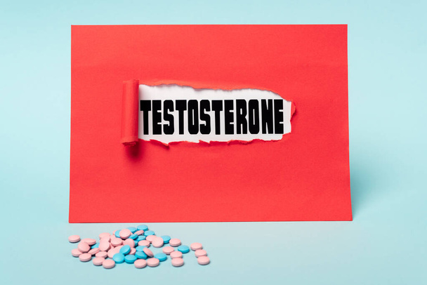 Mavi arka planda kırmızı kağıt deliğinde hormon hapları ve testosteron harfleri. - Fotoğraf, Görsel