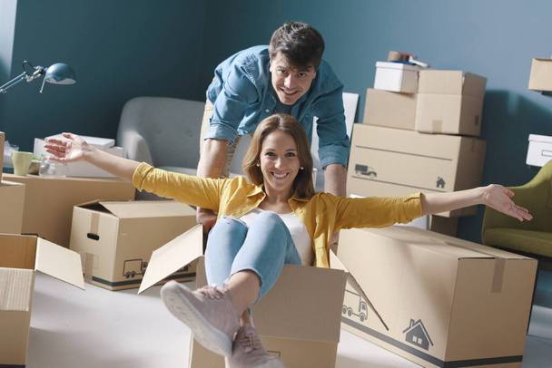 Cheerful happy young couple moving in their new house: she is sitting in a box and her boyfriend is pushing her - Φωτογραφία, εικόνα