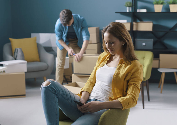 Woman sitting on a chair chatting with her phone during home relocation, her boyfriend is moving boxes in the background - Foto, Imagen