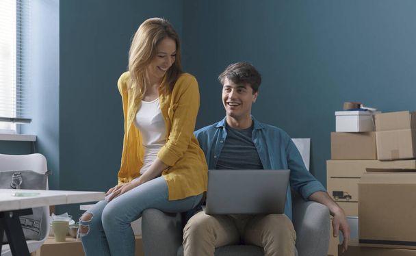 Happy young couple moving in their new home, they are sitting on the armchair together and connecting with their laptop, home relocation concept - Foto, Imagem