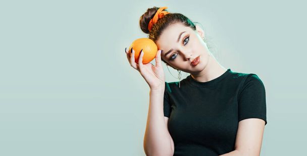 retrato de una hermosa joven con peinado de moño sosteniendo naranja cerca de la cabeza, retrato de moda en el fondo del estudio con luz verde
 - Foto, imagen