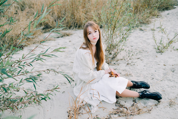 Портрет молодої дівчини в білій сукні, теплий плетений кардиган і чорне взуття серед сухого очерету і зелених кущів на березі річки з білим піском. - Фото, зображення