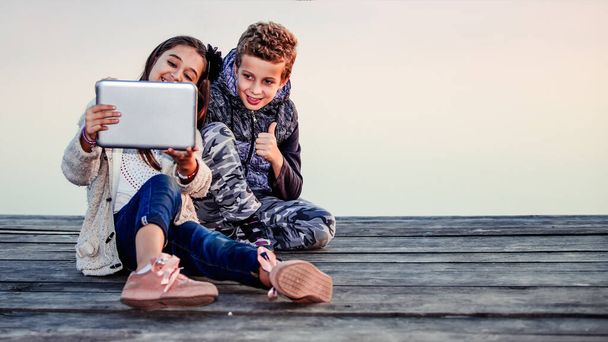 Two young cute little friends, boy and girl having fun playing on digital tablet sitting by the lake in the evening. Children celebrating success.  The boy is showing gesture thumb up.Friendship - Photo, Image