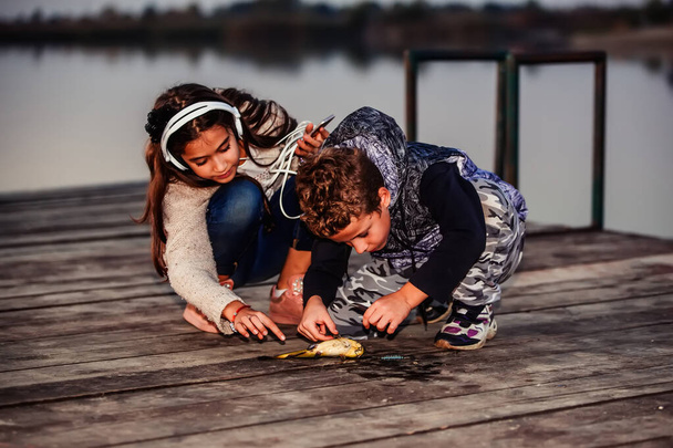 Dos jóvenes lindos amiguitos, niño y niña pescando en un lago por la noche. Un niño pequeño mostrando a su amigo un pez atrapado. Los niños están jugando. Amistad
. - Foto, Imagen