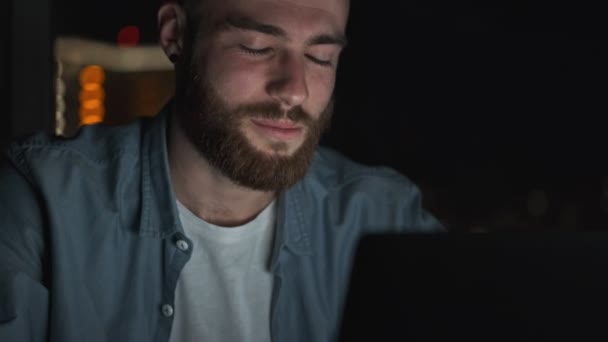A close-up view of a positive young man wearing casual clothes is talking to a video chat on his laptop computer at night time - Кадри, відео