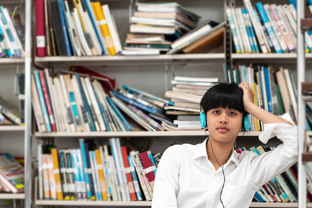 Adolescente joven con auriculares en la cabeza, sentada frente a la estantería del libro, con sensación probada, en la biblioteca, luz borrosa alrededor
 - Foto, imagen