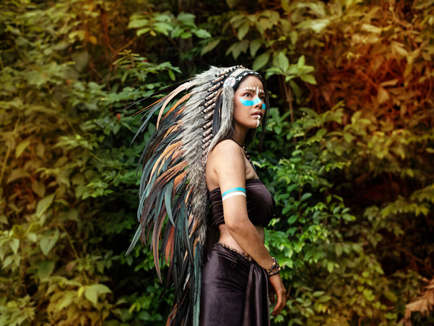 Η όμορφη γυναίκα με το καπέλο φτιαγμένο από φτερά πουλιών. βαμμένο μπλε χρώμα στο μάγουλό της, πορτρέτο του μοντέλου που θέτει, ζούγκλα στο δάσος, θολή φως γύρω - Φωτογραφία, εικόνα