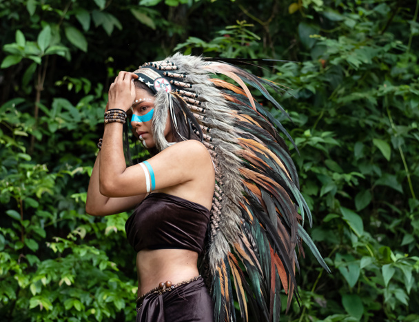 La belle femme soulevant coiffe toucher la main faite de plumes d'oiseaux. couleur bleue peinte sur sa joue, portrait de mannequin posant, jungle dans la forêt, lumière floue autour
 - Photo, image