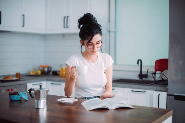 Beautiful young woman reading book in the kitchen - Фото, изображение