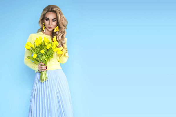 Spring fashion Woman. Beautiful model with colorful clothes, holding a bouquet of spring flowers posing over blue background . Fashion photo. Easter concept. - Photo, Image