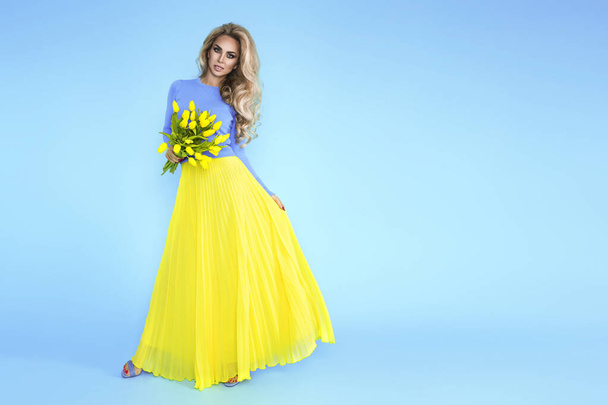 Frühlingsmode Frau. Schöne Modell mit bunten Kleidern, hält einen Strauß Frühlingsblumen posiert über blauem Hintergrund. Modefoto. Osterkonzept. - Foto, Bild