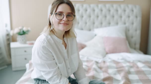 Beautiful girl in glasses smiling sitting on bed in bedroom apartment looking at camera dressed in white shirt, woman mother in a good mood portrait of an independent and confident young girl. - 映像、動画