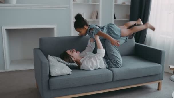 Friendly beautiful family healthy young mother lifting cool child daughter up playing plane on sofa at apartment, funny kid girl having fun together fly in mothers arm practice acro pair yoga bonding - Video, Çekim