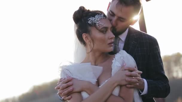 Bearded groom hugs from behind and kisses his beloved bride against the backdrop of the bright sun in a deserted field. Walk in the fall on the nature of the wedding couple after the wedding ceremony. - Footage, Video