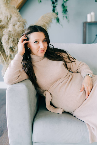 portrait of a young beautiful pregnant woman in a beige dress sitting on a gray sofa - Photo, Image